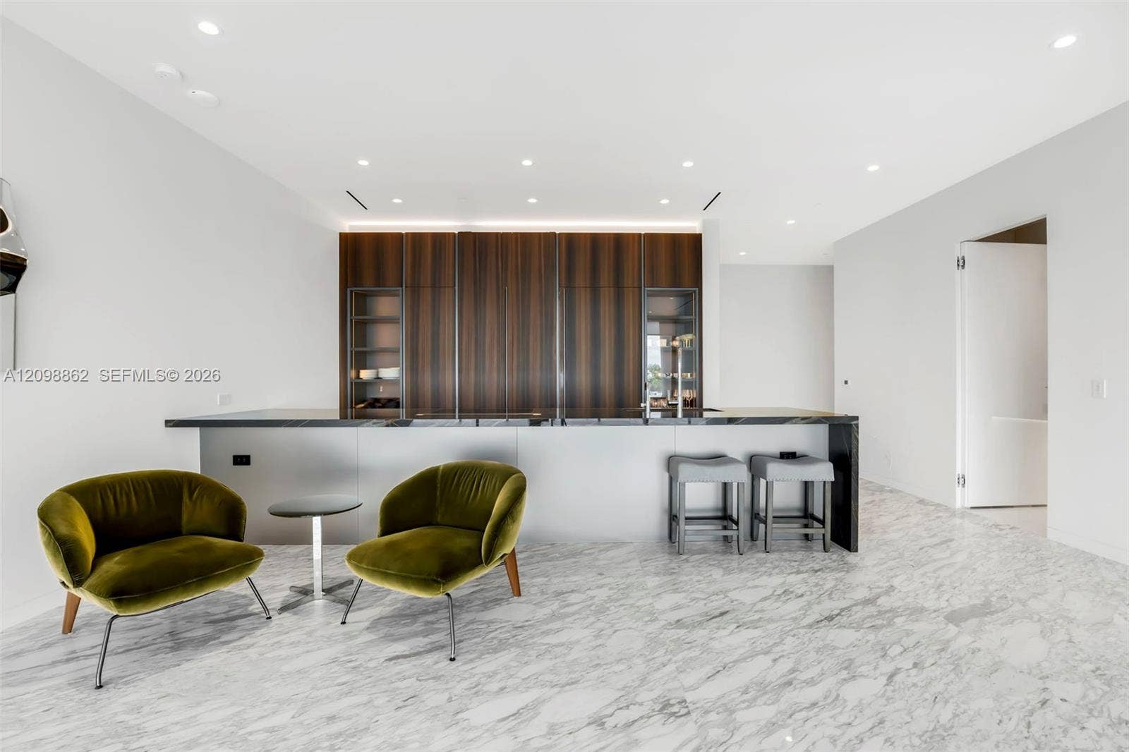 Modern kitchen featuring full-height wood-finish cabinetry, sleek stone-surfaced island, recessed lighting, and continuous stone-patterned flooring