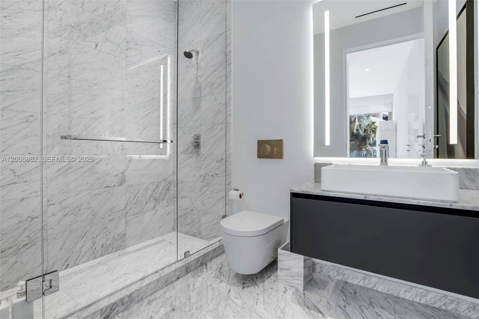 Full-height stone wall tiling, glass-enclosed walk-in shower, wall-mounted commode with metallic flush plate, floating vanity with rectangular vessel sink, and integrated LED-backlit mirror