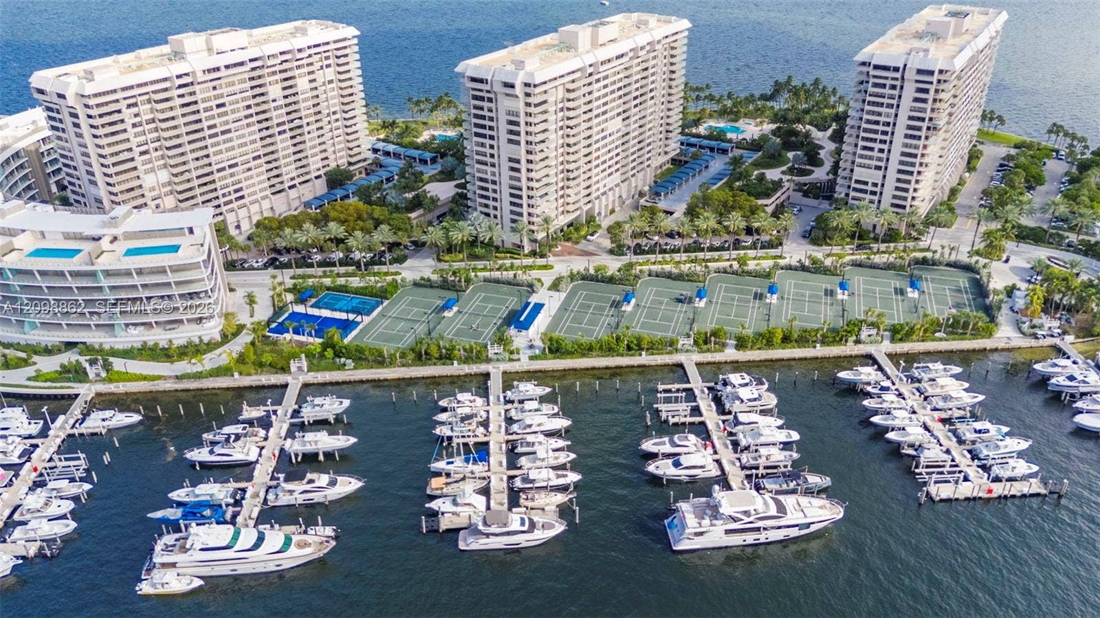 Aerial perspective of a waterfront complex featuring a private deep-water marina, multiple tennis courts, swimming pools, and landscaped grounds