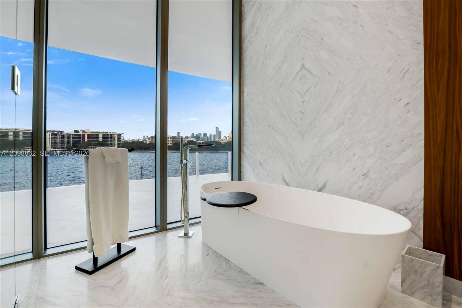 Freestanding soaking tub with floor-mounted chrome faucet, stone-surfaced wall, floor-to-ceiling glass paneling, and stone-finish flooring