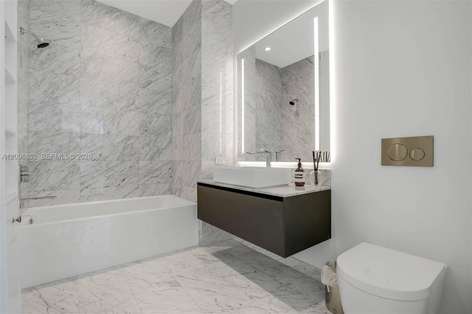Full-height natural stone tiling in shower and tub surround, floating vanity with rectangular vessel sink, wall-mounted faucet, integrated LED-lit vanity mirror, and wall-mounted dual-flush toilet