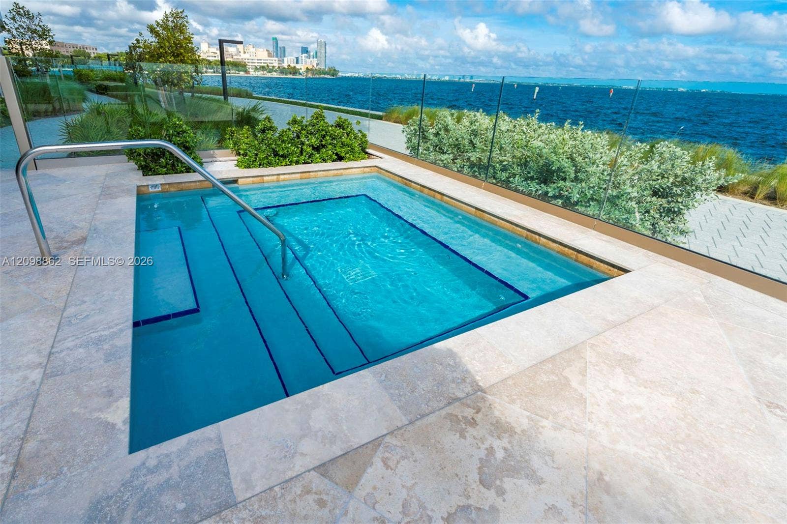 In-ground spa with integrated staircase and stainless steel handrails, surrounded by stone paver decking and a glass perimeter railing, overlooking a coastal waterfront