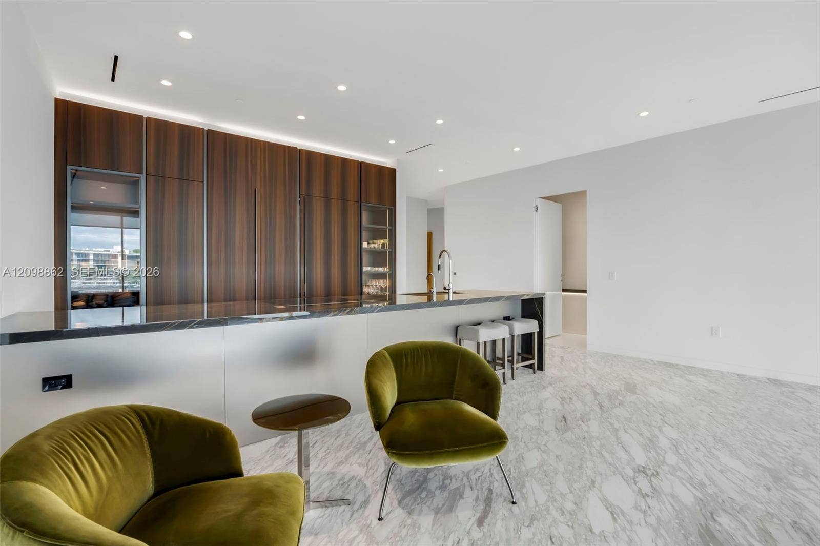 Modern kitchen featuring floor-to-ceiling wood-finish cabinetry, a large stone-surfaced island with integrated sink, recessed ceiling lighting, and light-toned stone flooring