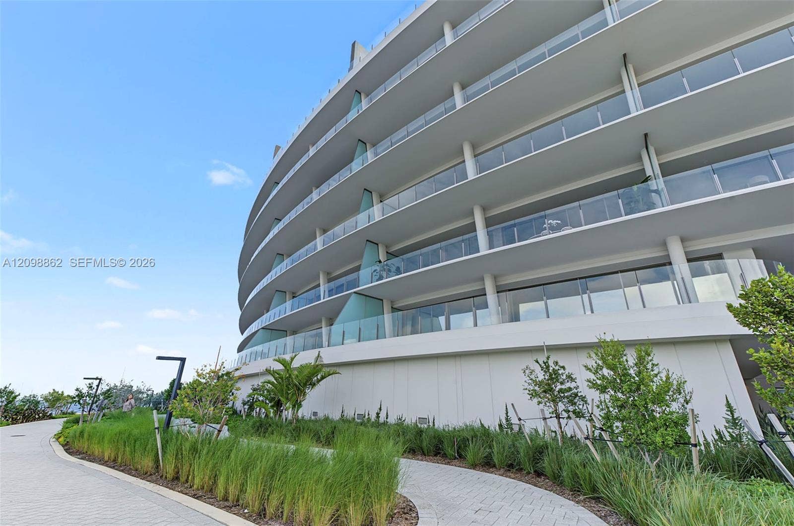 Curved multi-story exterior featuring tiered balconies, glass railings, concrete structural columns, and landscaped walking paths with native grasses