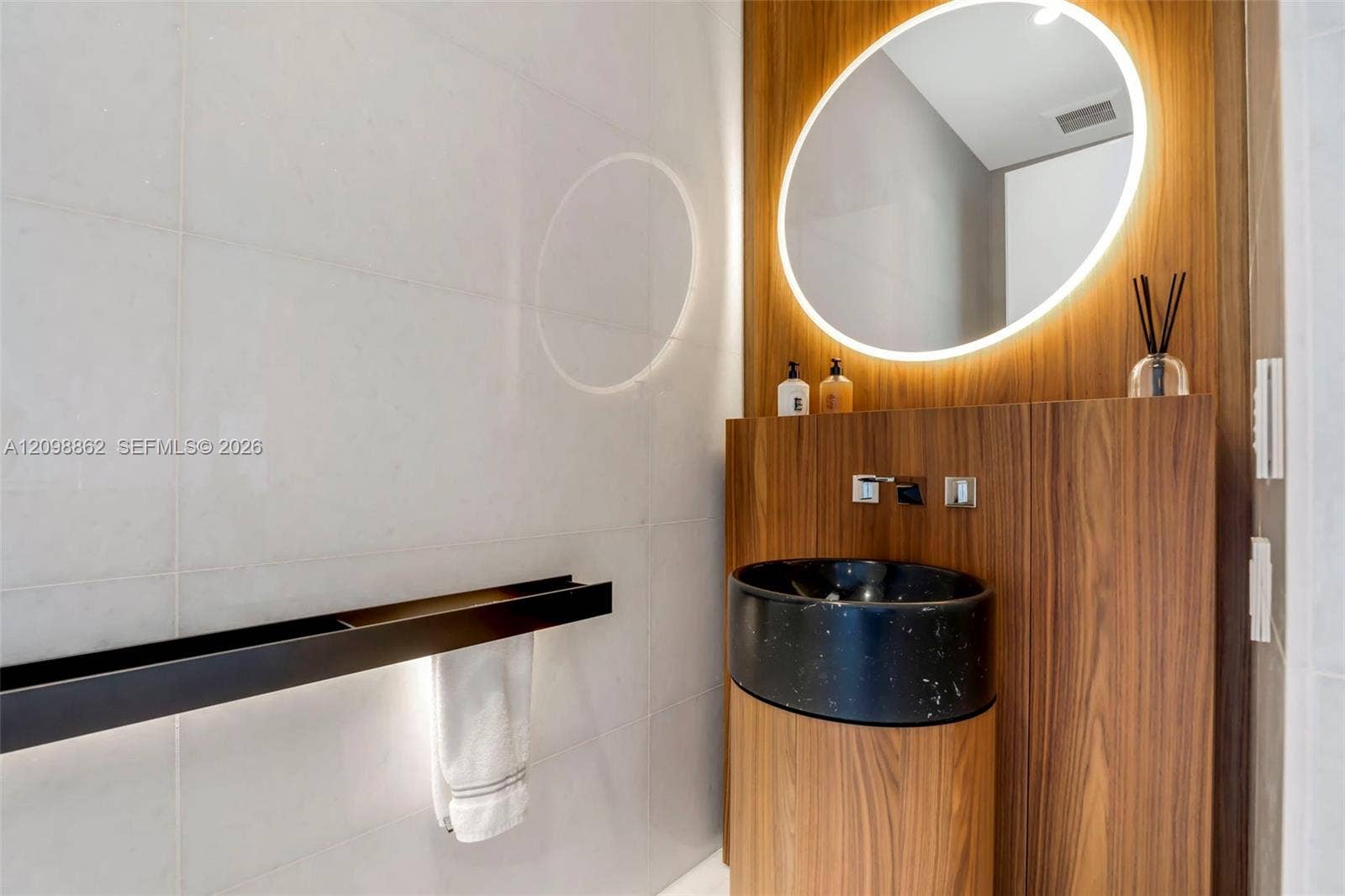 Wall-mounted wood-finish vanity with a circular stone vessel sink, integrated LED-backlit mirror, large-format tile wall cladding, and matte black linear fixture