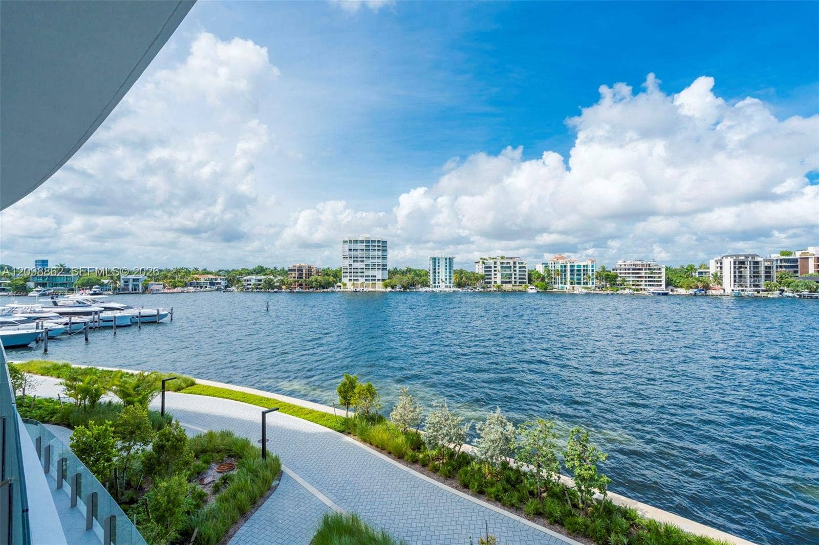 Covered balcony featuring a paved waterfront promenade, manicured landscaping, and direct water frontage