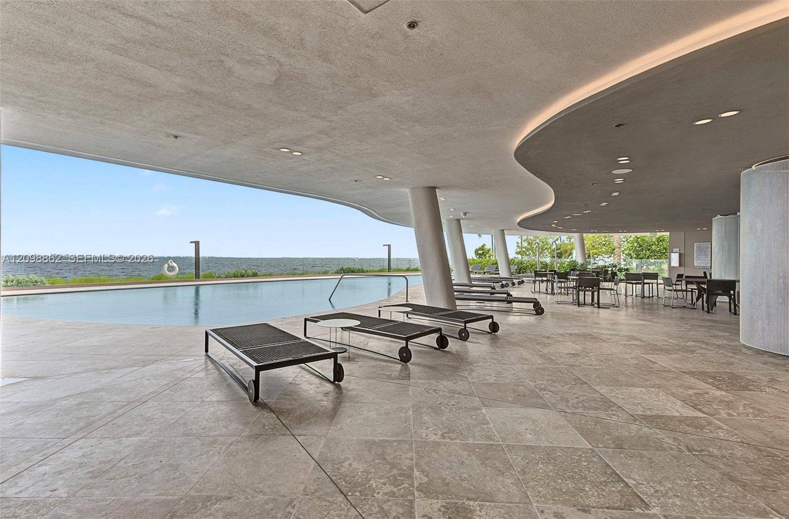 Covered poolside terrace featuring expansive stone flooring, a curved architectural ceiling with recessed lighting, structural support columns, and an infinity-edge swimming pool overlooking the water