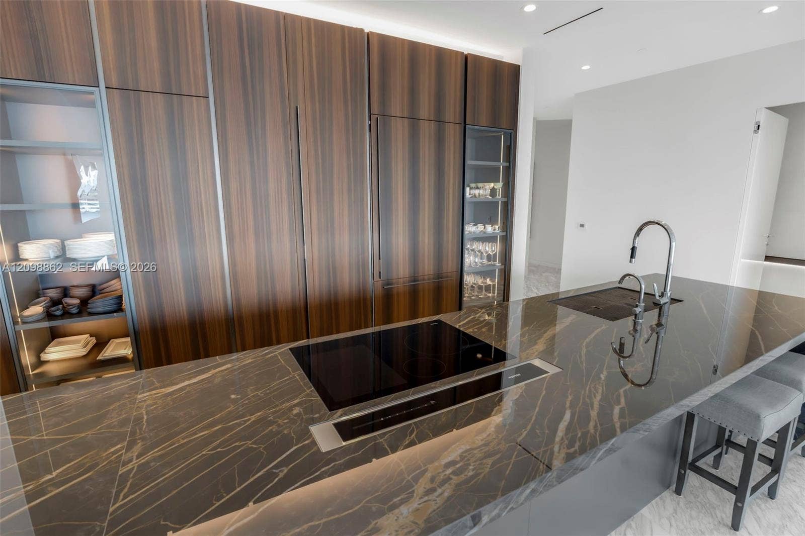 Floor-to-ceiling wood-finish cabinetry with integrated appliances, stone-surface kitchen island, induction cooktop, built-in downdraft vent, and illuminated glass display shelving