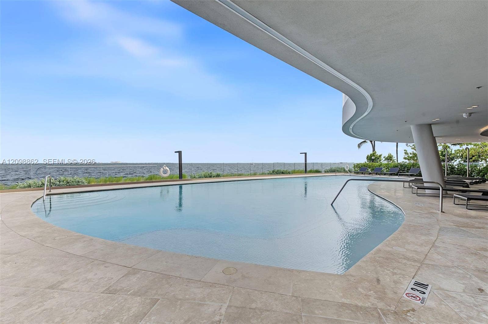 Curved-edge infinity pool featuring stone decking, glass railings, and a covered patio area