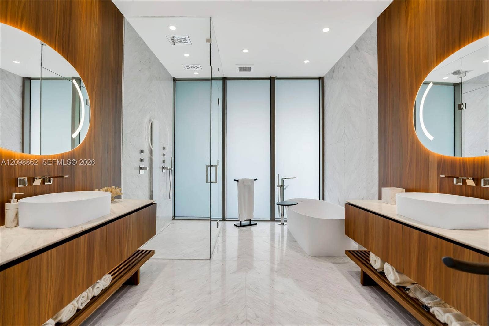 Dual floating wood-finish vanities with stone countertops, vessel sinks, and integrated backlit oval mirrors, complemented by a stone-tiled floor, a glass-enclosed shower, and a freestanding soaking tub