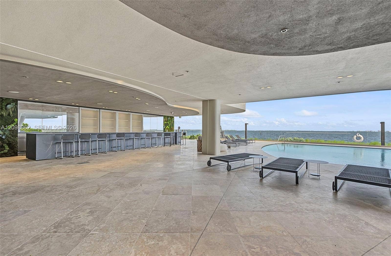 Covered outdoor lounge featuring stone tile flooring, a custom-built bar with integrated shelving, recessed ceiling lighting, and an infinity-edge pool