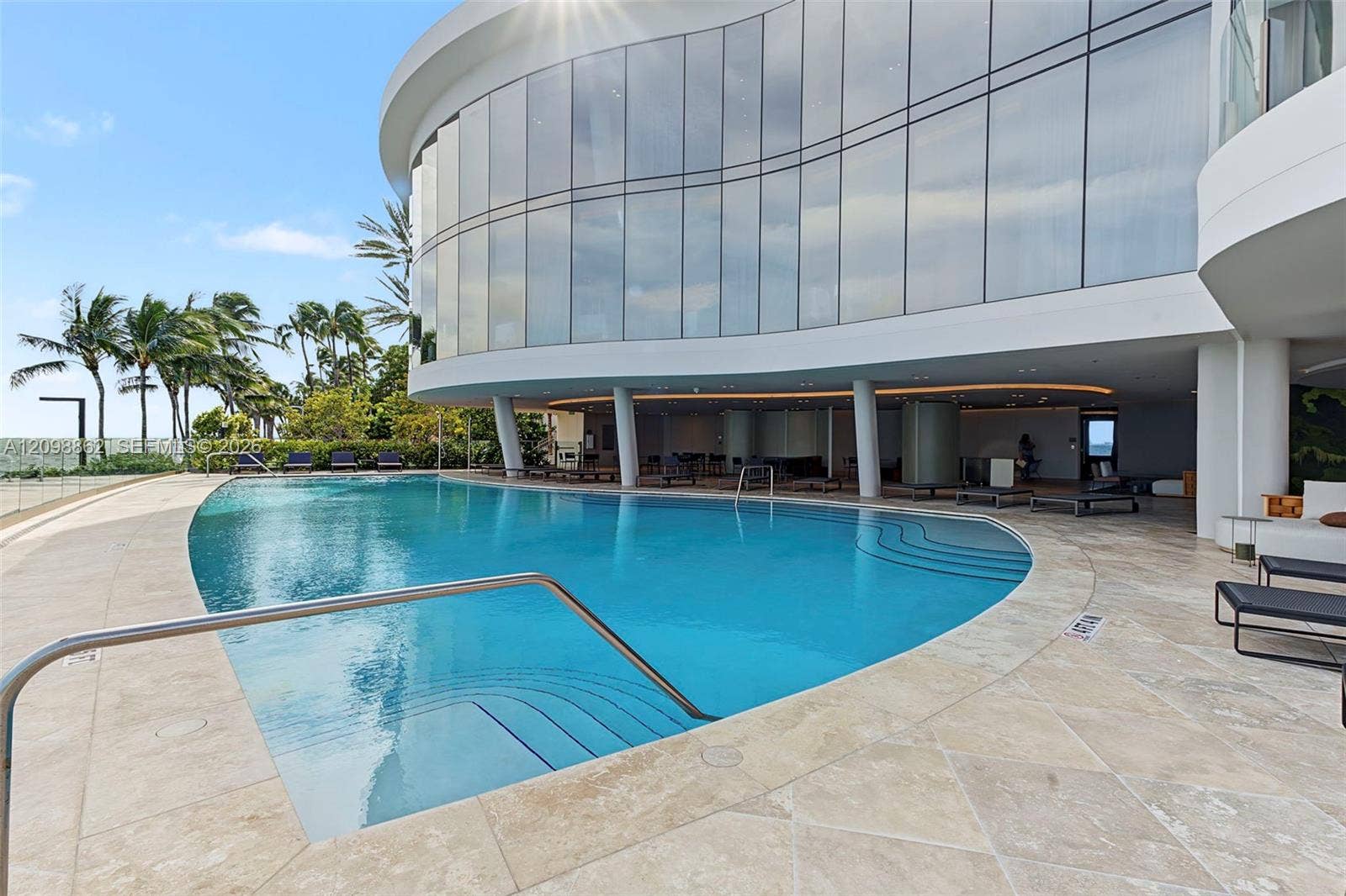 Curved structural exterior featuring floor-to-ceiling glass wall panels, expansive stone-tile pool deck, and custom-shaped swimming pool with built-in stairs