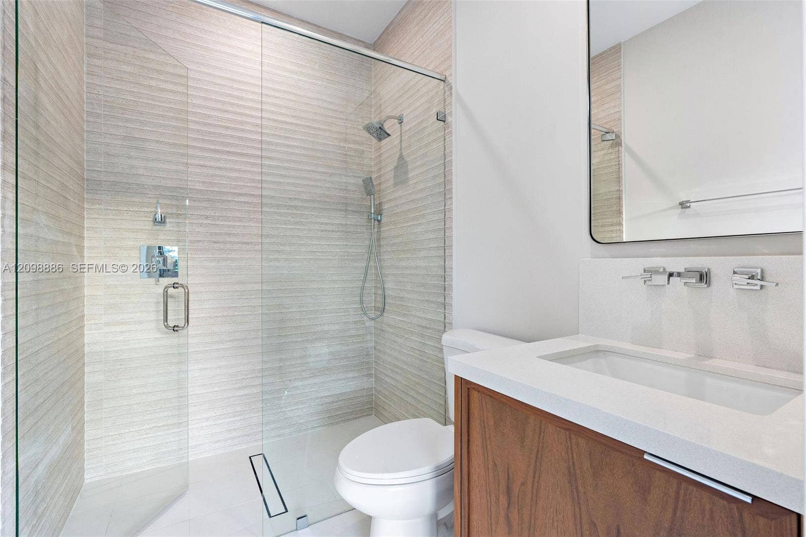 Walk-in shower featuring textured wall tile, glass enclosure, and rainfall showerhead, paired with a wood-finish vanity, solid-surface countertop, and wall-mounted faucet
