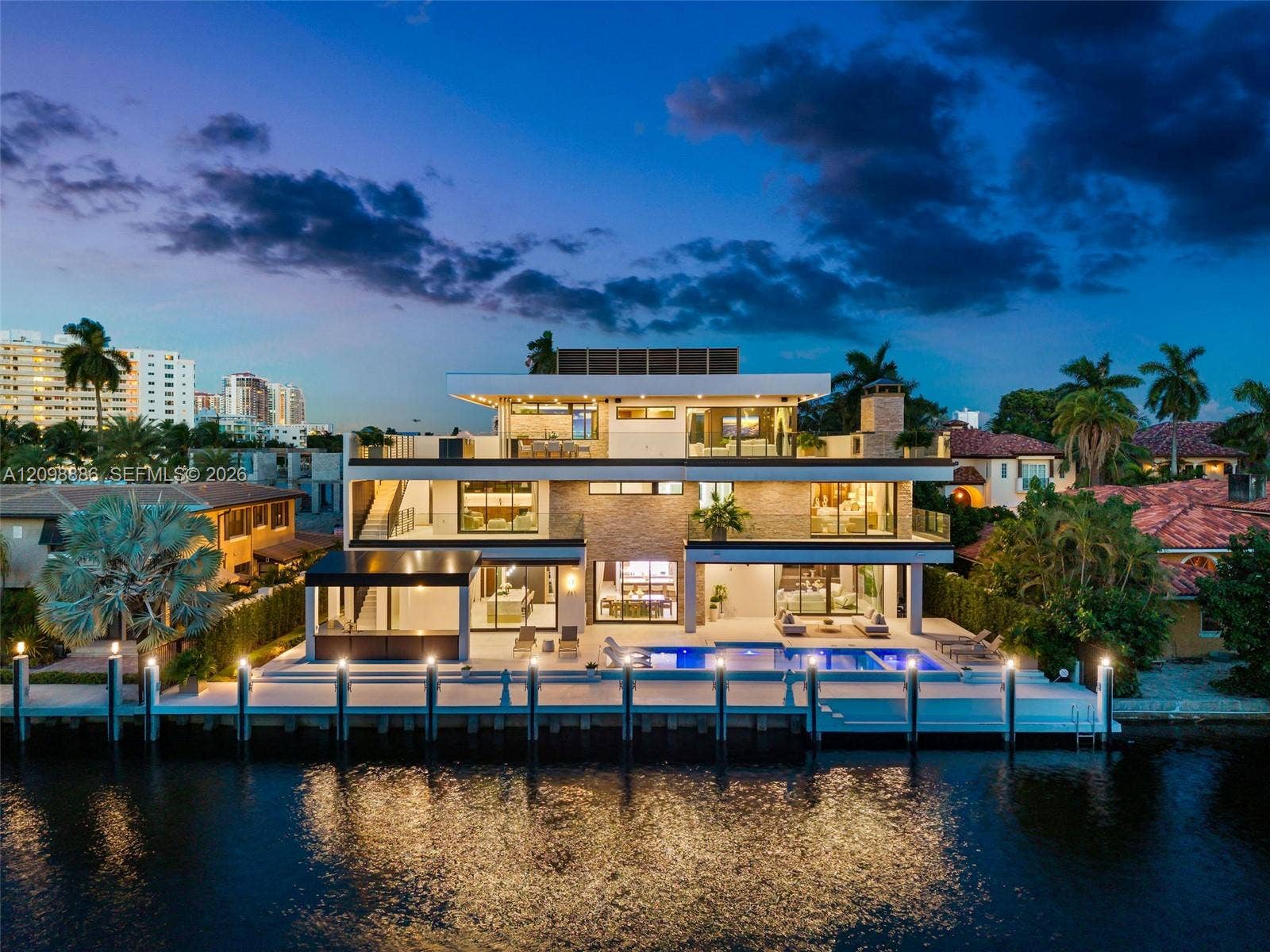 Three-story modern waterfront residence featuring a private boat dock, multi-level terraces with glass railings, stone veneer accents, and a rectangular swimming pool