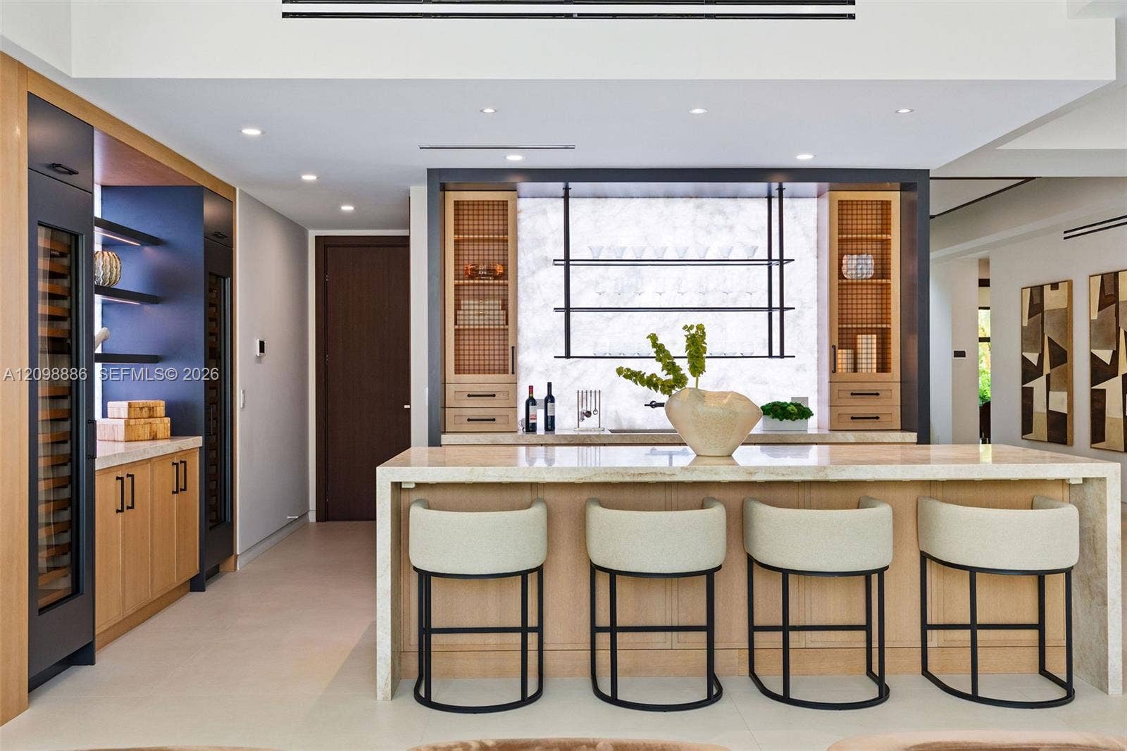 Custom bar area featuring stone slab backsplash, integrated wine refrigeration, built-in light wood cabinetry, stone-topped island with waterfall edge, and tiered floating shelving