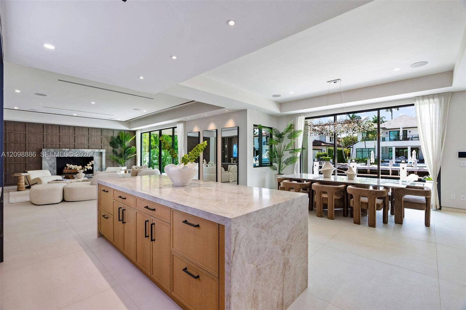 Large kitchen island featuring a waterfall stone countertop and light wood-finish cabinetry with matte black hardware, stone tile flooring, a floor-to-ceiling stone fireplace surround, and expansive sliding glass doors