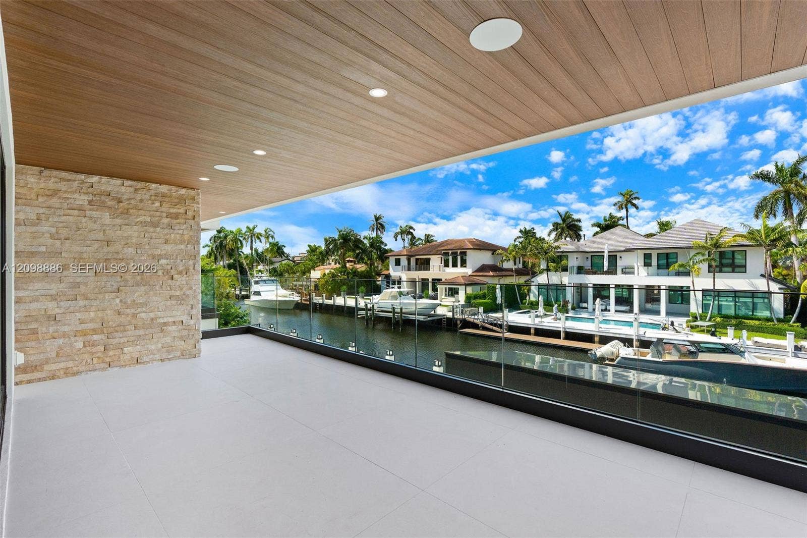 Covered balcony featuring wood-finish ceiling paneling, stacked stone accent wall, large-format tile flooring, and a frameless glass railing overlooking the canal