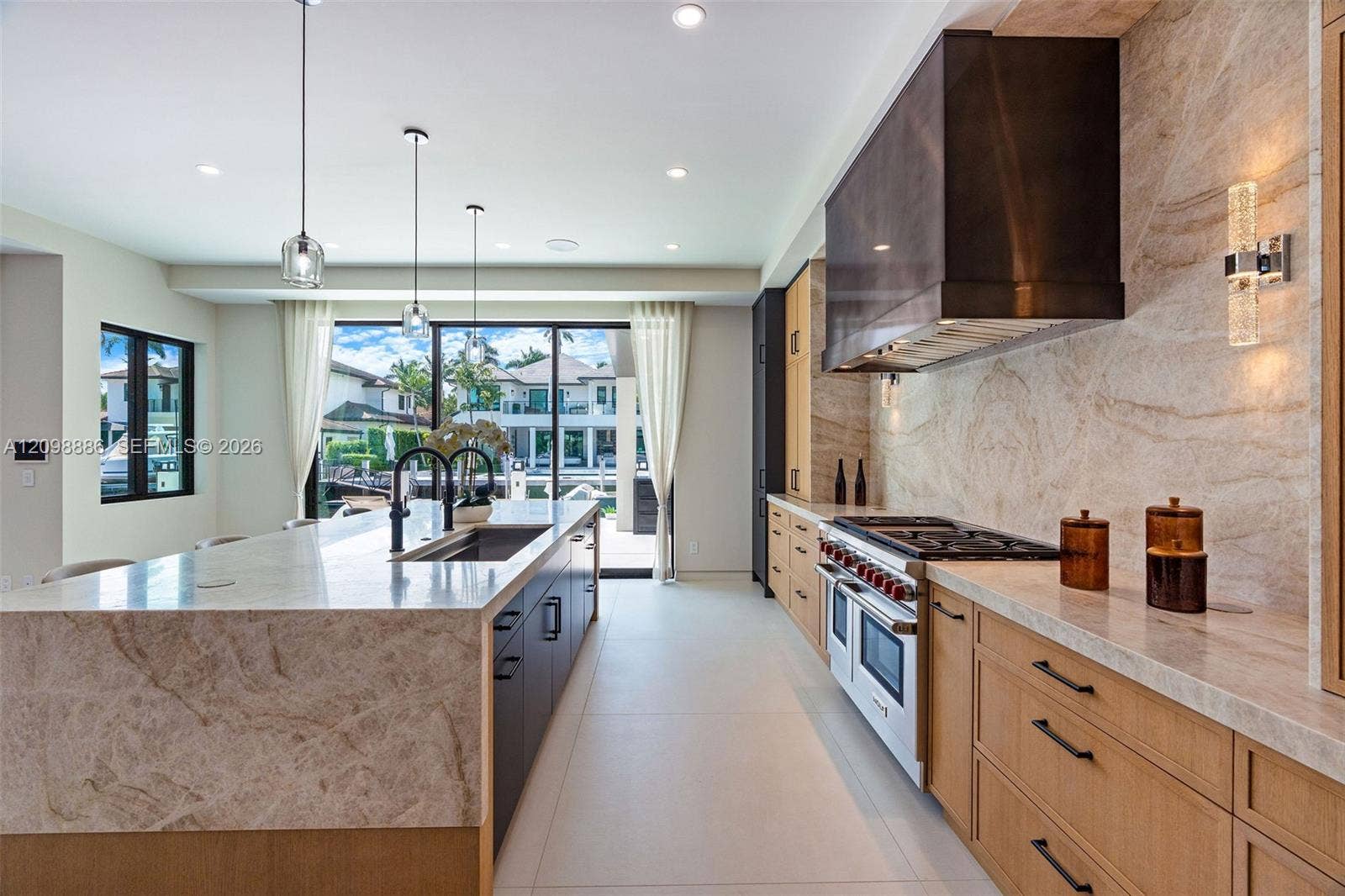 Kitchen featuring custom wood cabinetry, natural stone countertops and backsplash, professional-grade range, custom metal range hood, and large center island with integrated sink