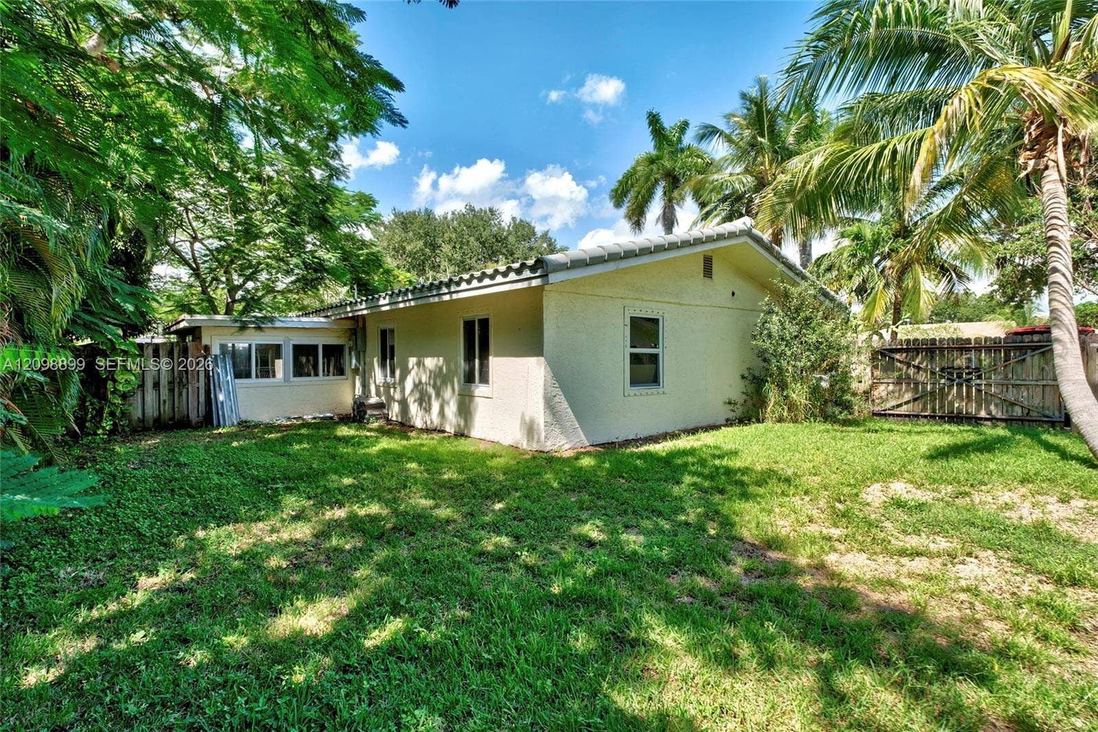 1176 SW 4th Street, Boca Raton, FL 33486 Stucco exterior residence featuring a tiled roof, expansive lawn, mature perimeter trees, and a wooden privacy fence