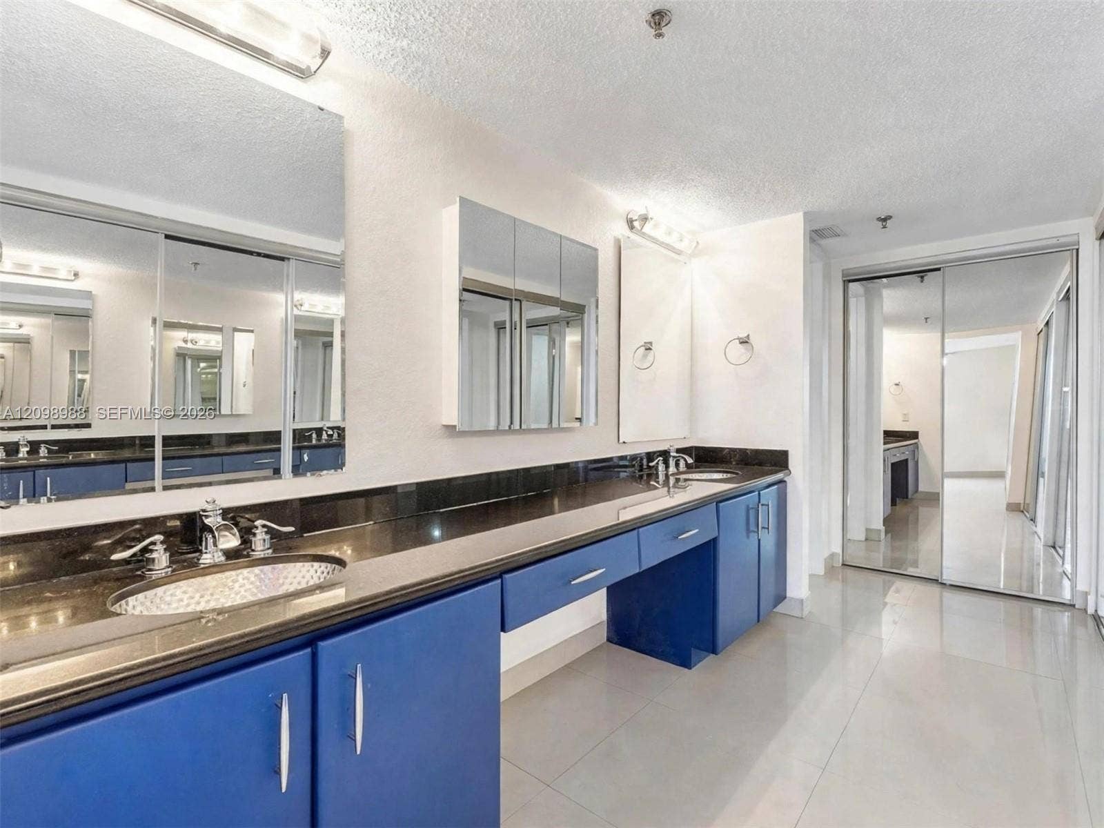 Extended dual-sink vanity featuring blue cabinetry, polished dark countertops, integrated oval basins, chrome-finish fixtures, and full-width mirror wall