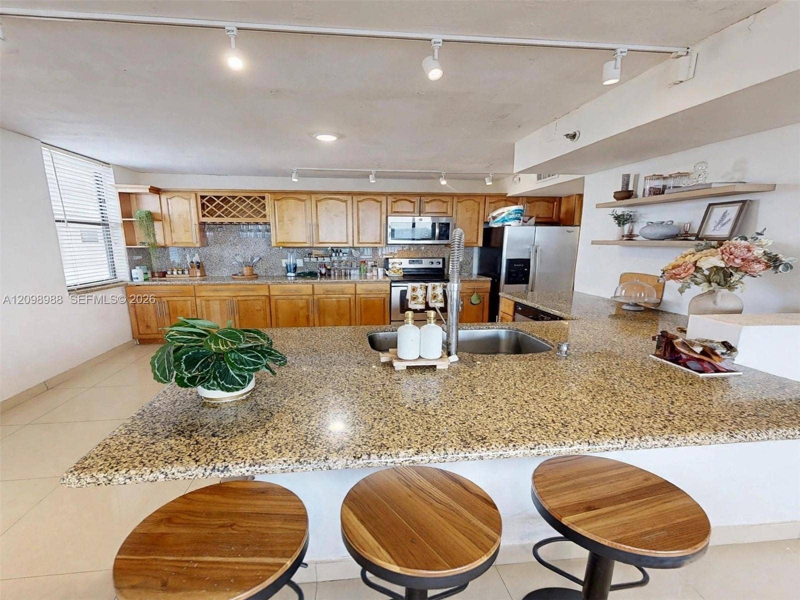 Kitchen featuring a stone-surfaced island with an integrated sink, wooden cabinetry, track lighting, and built-in floating shelves
