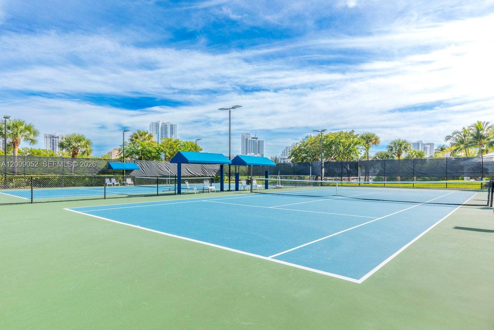 1516 Breakwater Terrace, Hollywood, FL 33019 Hard-surface tennis courts featuring perimeter mesh fencing, blue sunshades, integrated court lighting, and mature landscaping