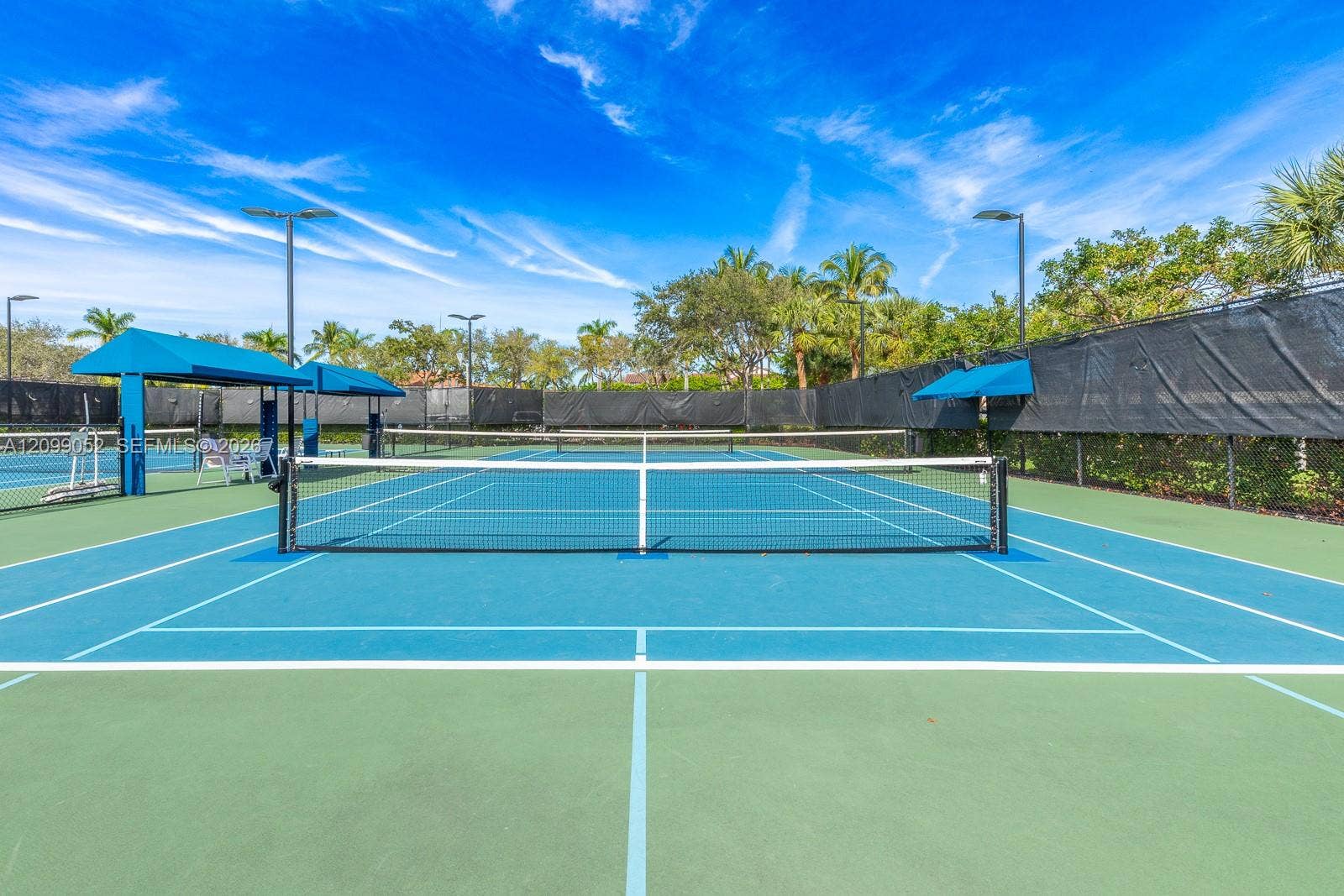 1516 Breakwater Terrace, Hollywood, FL 33019 Hard-court tennis facility featuring blue and green surface, permanent net, protective perimeter fencing, pole-mounted lighting, and integrated shaded structures