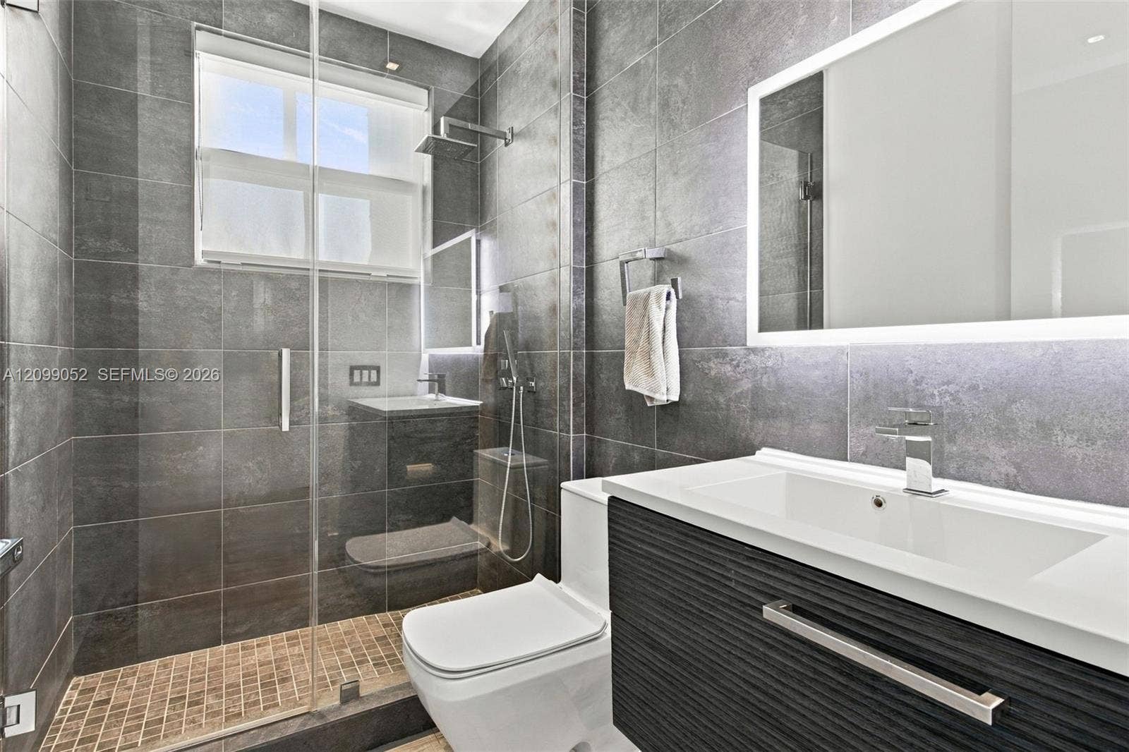1516 Breakwater Terrace, Hollywood, FL 33019 Bathroom featuring large-format dark grey wall tile, a glass-enclosed shower with a rainfall showerhead, a floating vanity with a textured cabinet finish, and a rectangular undermount sink with a backlit mirror