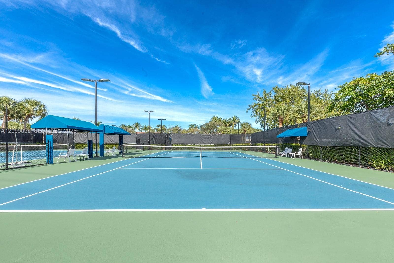 1516 Breakwater Terrace, Hollywood, FL 33019 Hard-court tennis facility featuring blue and green surface markings, perimeter privacy fencing, overhead lighting, and built-in shaded seating areas