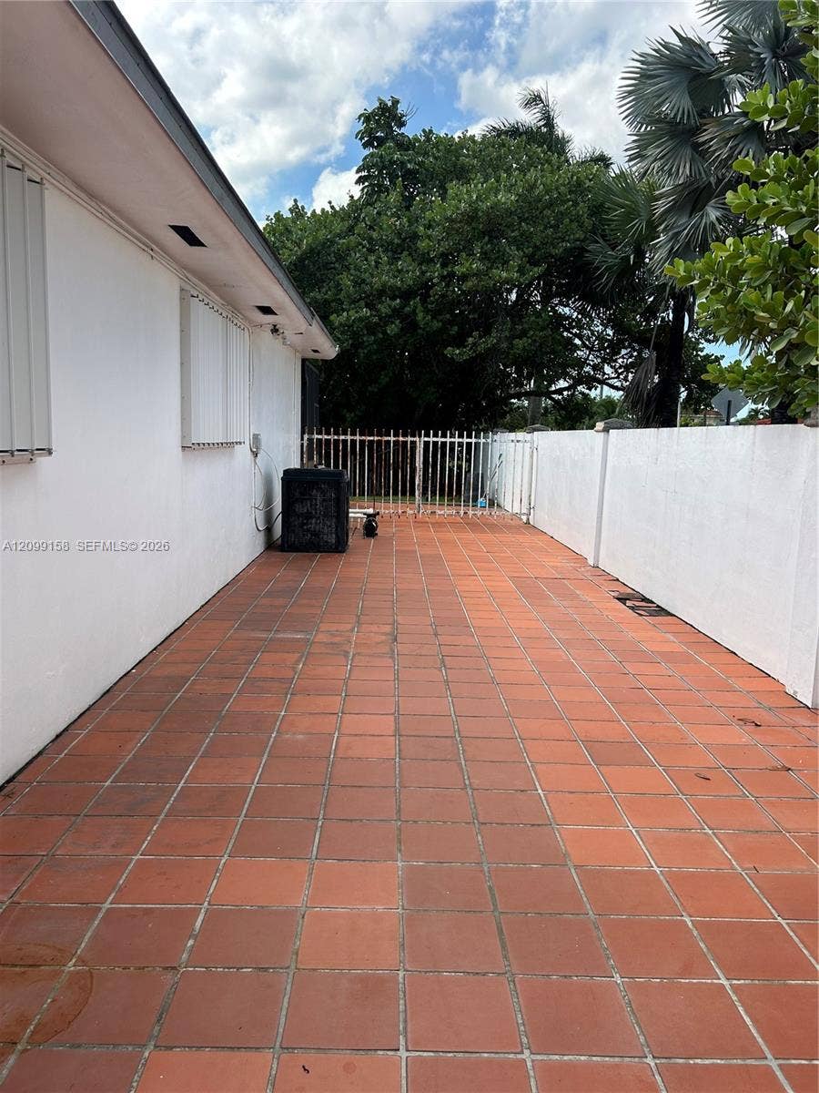 1490 Lenape Drive, Miami Springs, FL 33166 Exterior side patio featuring terracotta tile flooring, enclosed by a white masonry privacy wall and gated metal fencing