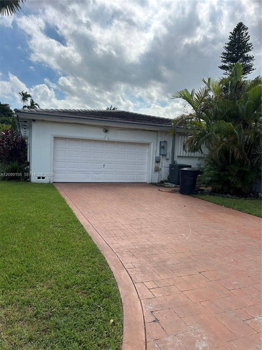 1490 Lenape Drive, Miami Springs, FL 33166 Attached two-car garage with a white roll-up door, complemented by a textured paved driveway and manicured lawn