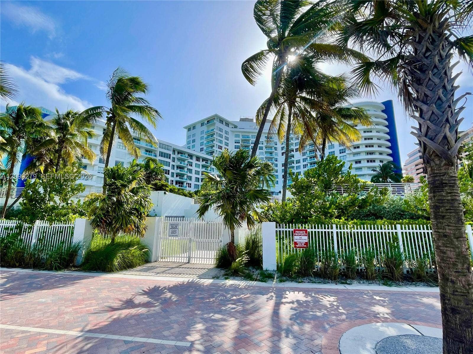 5151 Collins Avenue, #921, Miami Beach, Miami Beach, FL 33140 High-rise building with rounded architectural detailing, white metal security fencing, paved brick pedestrian path, and mature palm trees