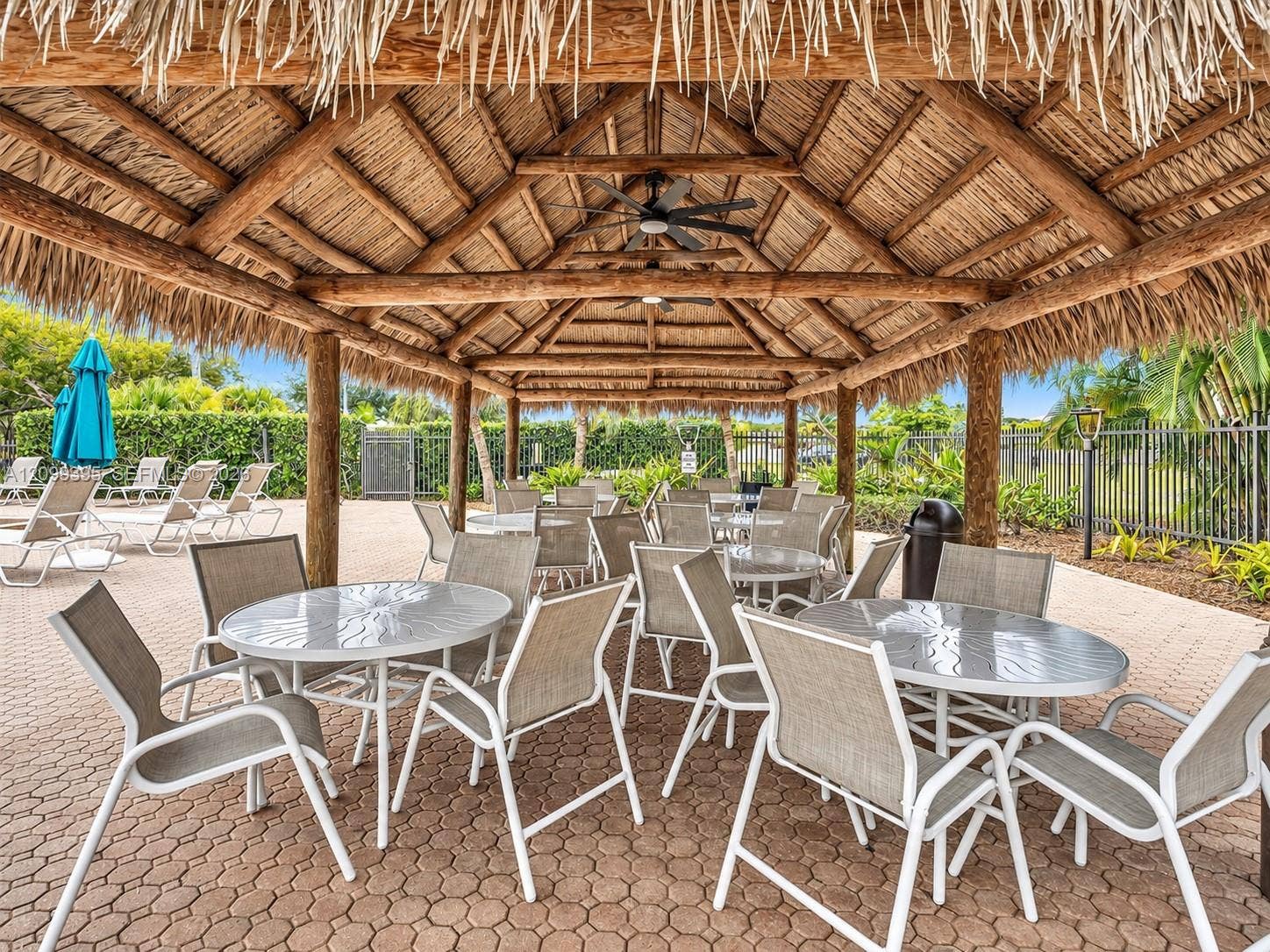 Outdoor pavilion featuring a thatched roof, exposed timber support beams, and ceiling-mounted fans over paved hardscaping