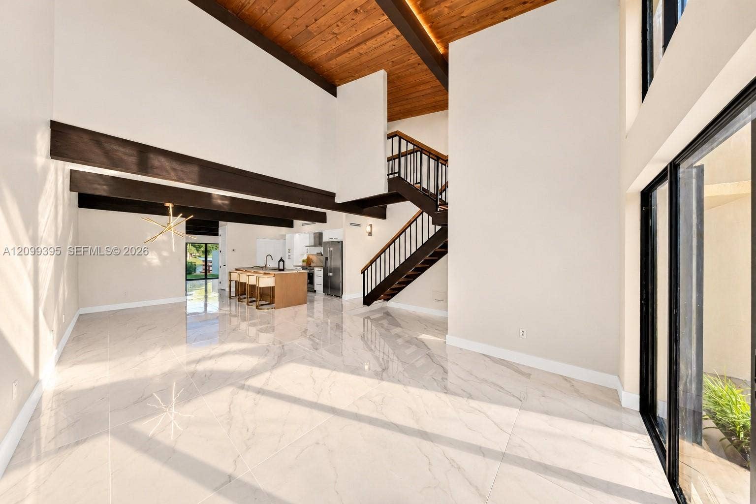 High-ceiling interior featuring a cedar wood-plank ceiling with exposed structural beams, large-format tile flooring, a geometric multi-arm light fixture, and a floating staircase with bronze metal railings