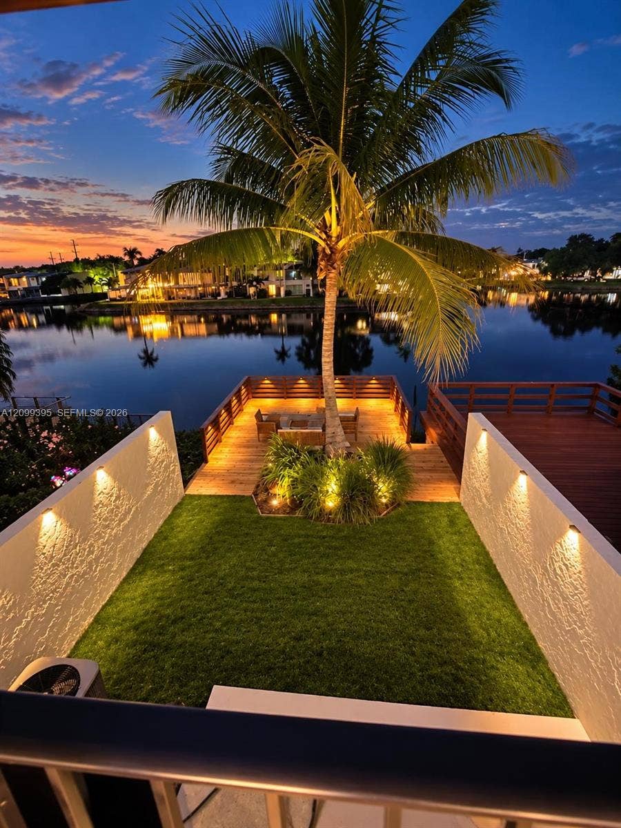 Waterfront property featuring a manicured lawn, wood-plank deck, integrated wall-mounted accent lighting, and a mature palm tree