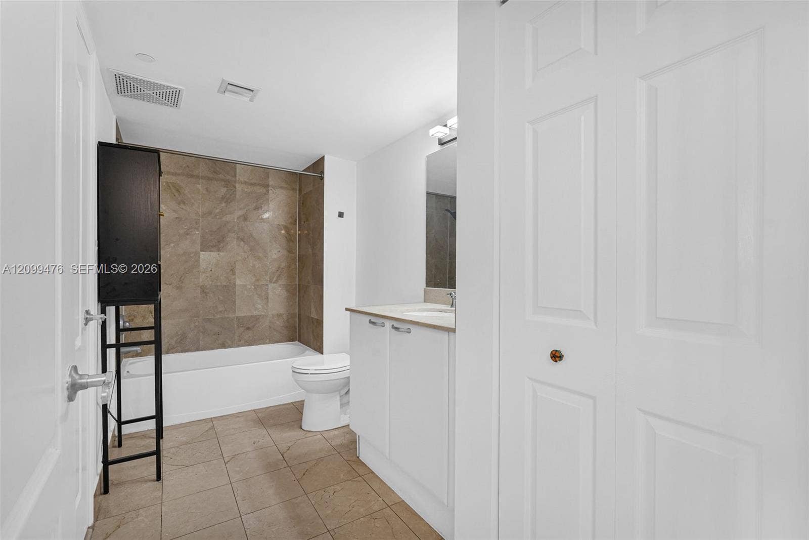 Bathroom featuring a white vanity cabinet with a solid-surface countertop, tiled tub surround, neutral-toned floor tile, and a multi-panel door closet