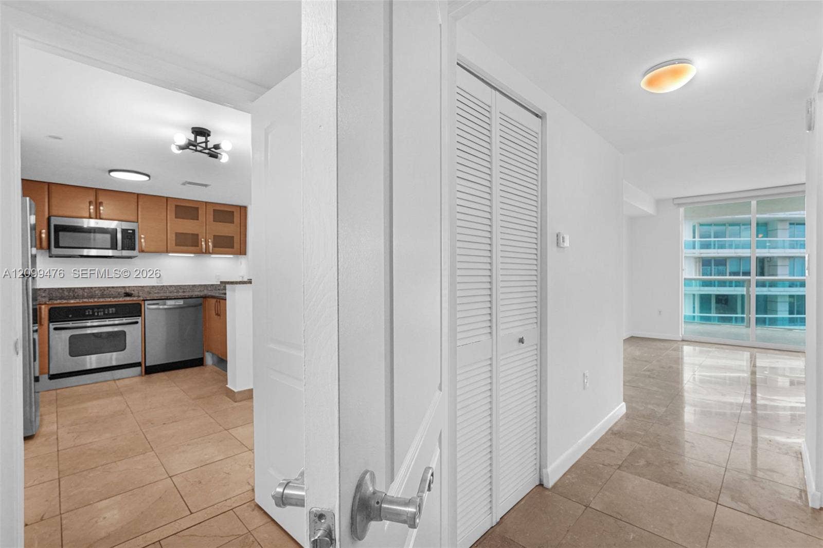 Kitchen featuring wood cabinetry, stainless steel appliances, and stone-finish countertops, paired with uniform tile flooring, louvered closet doors, and glass-paneled sliding balcony doors
