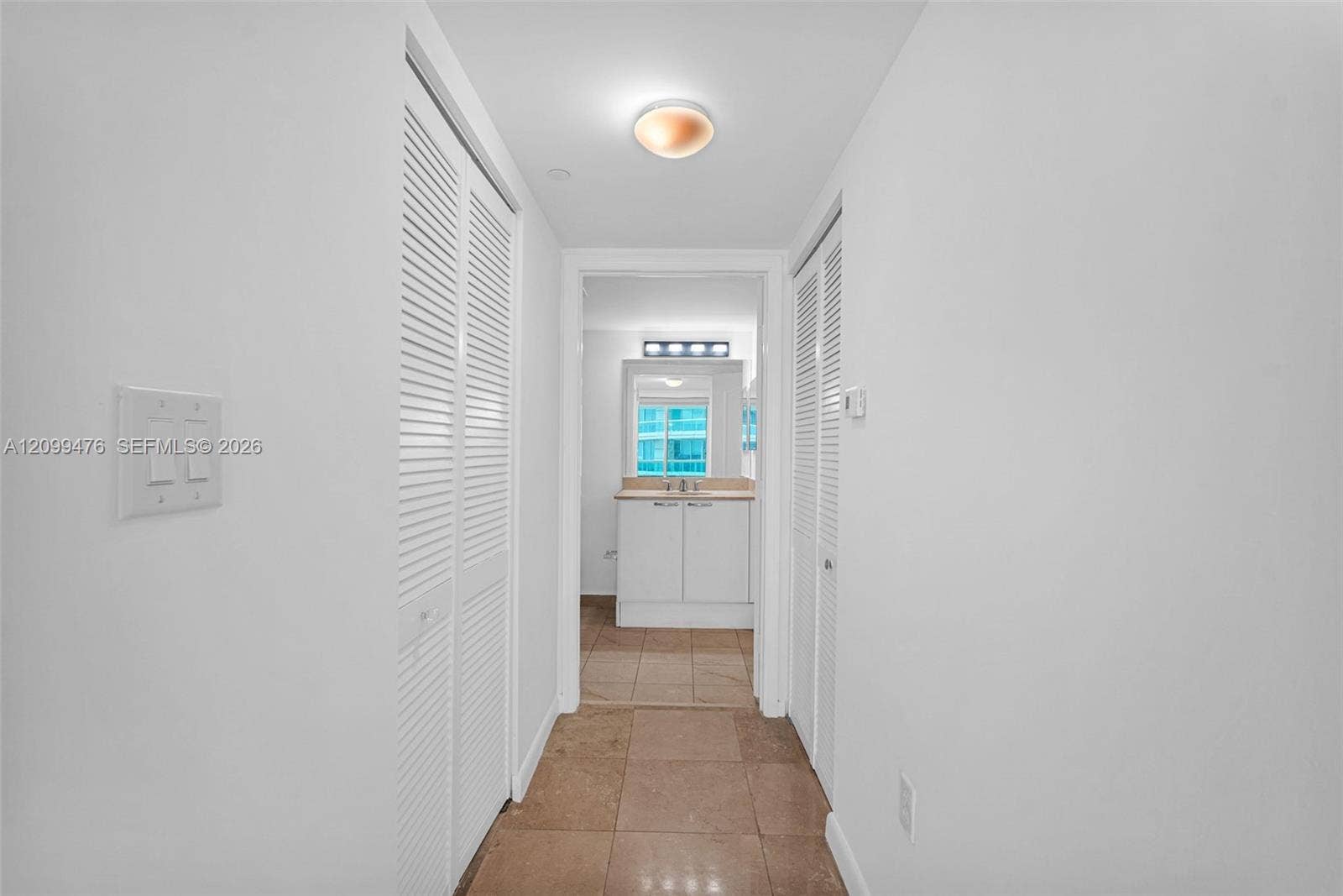 Hallway featuring dual sets of white louvered-door closets, neutral-toned tile flooring, and a flush-mount ceiling light fixture
