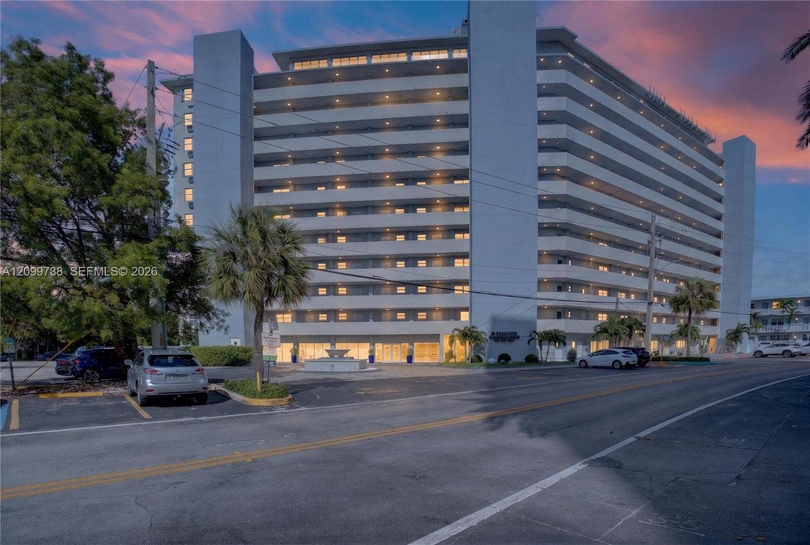 Multi-story residential building featuring wrap-around private balconies, a ground-level decorative fountain, and paved off-street parking