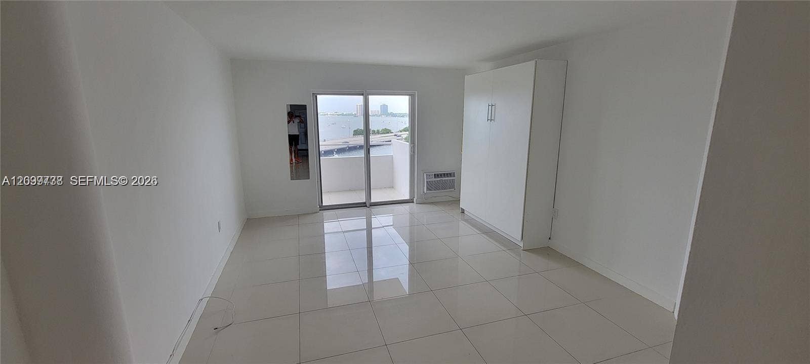 Neutral-toned interior featuring large-format tile flooring, built-in floor-to-ceiling cabinetry, and sliding glass doors opening to a balcony with waterfront scenery