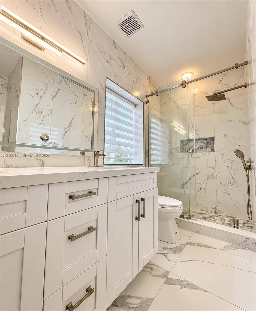 White vanity with stone-finish countertop, floor-to-ceiling stone-finish wall tile, walk-in glass-enclosed shower featuring a rainfall showerhead, and integrated wall niche