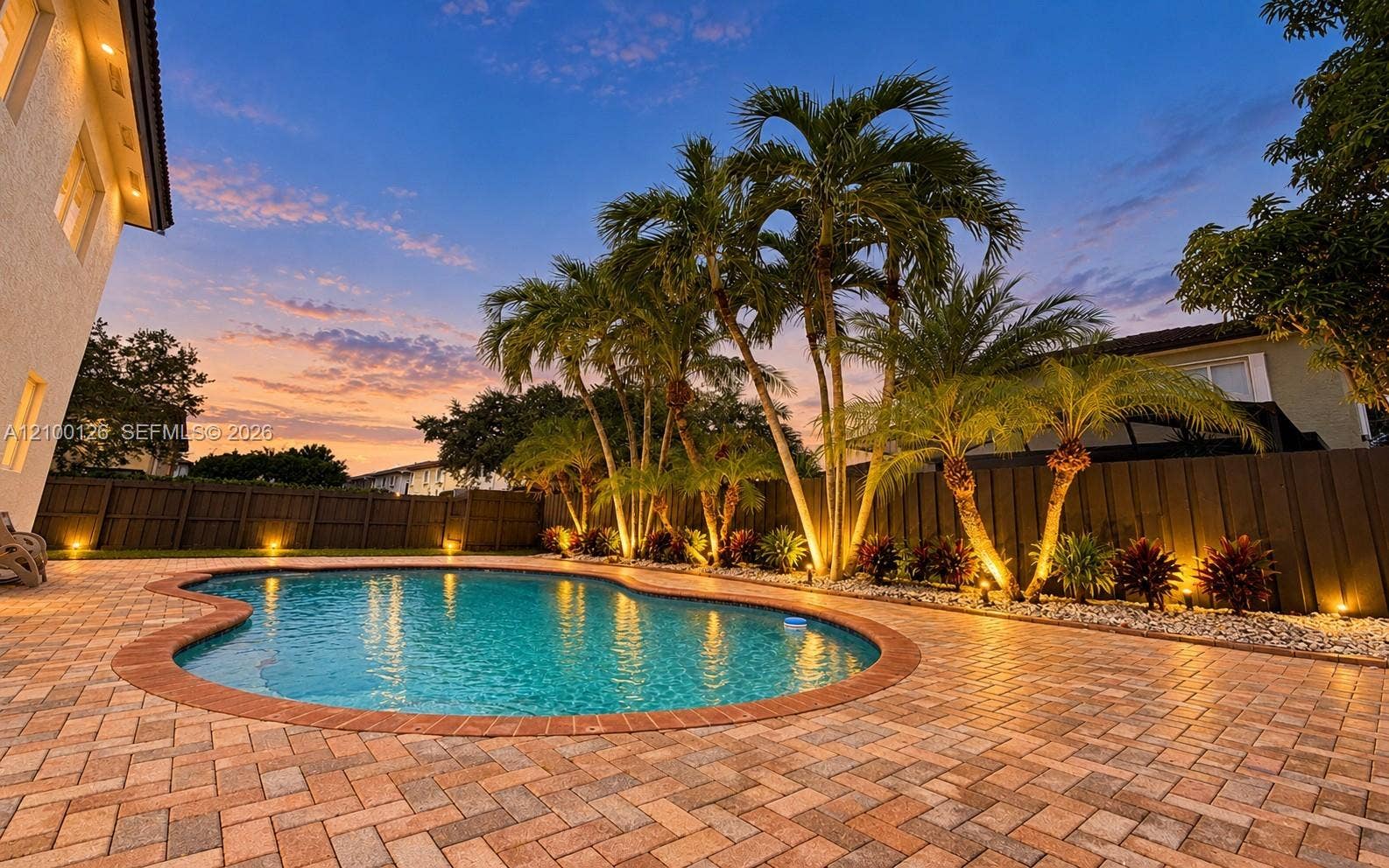 Custom-shaped swimming pool with brick-style paver decking, enclosed by privacy fencing, mature palm trees, and incorporated landscape lighting