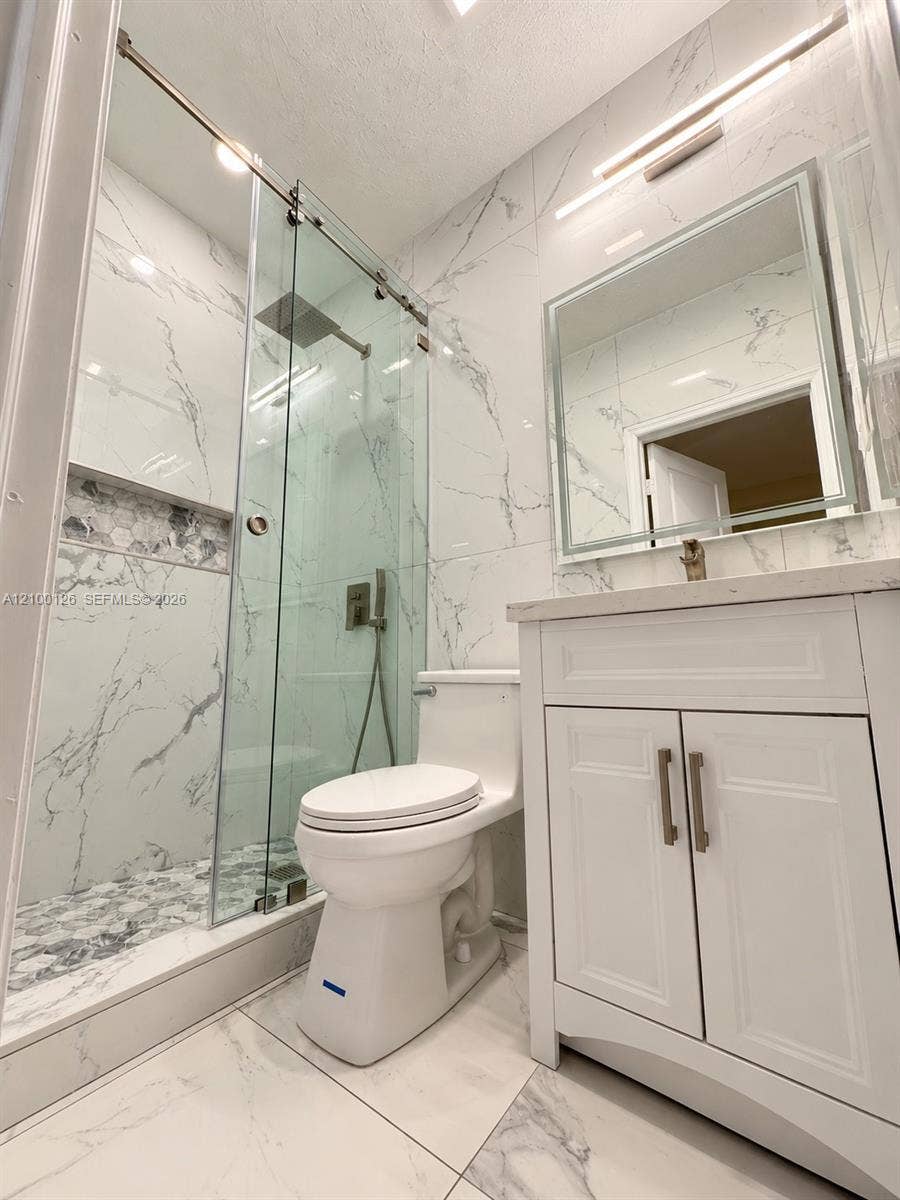 Full-height stone-finish wall tiling, glass-enclosed shower with a rainfall showerhead, white vanity with stone-finish countertop, backlit mirror, and integrated shower wall niche