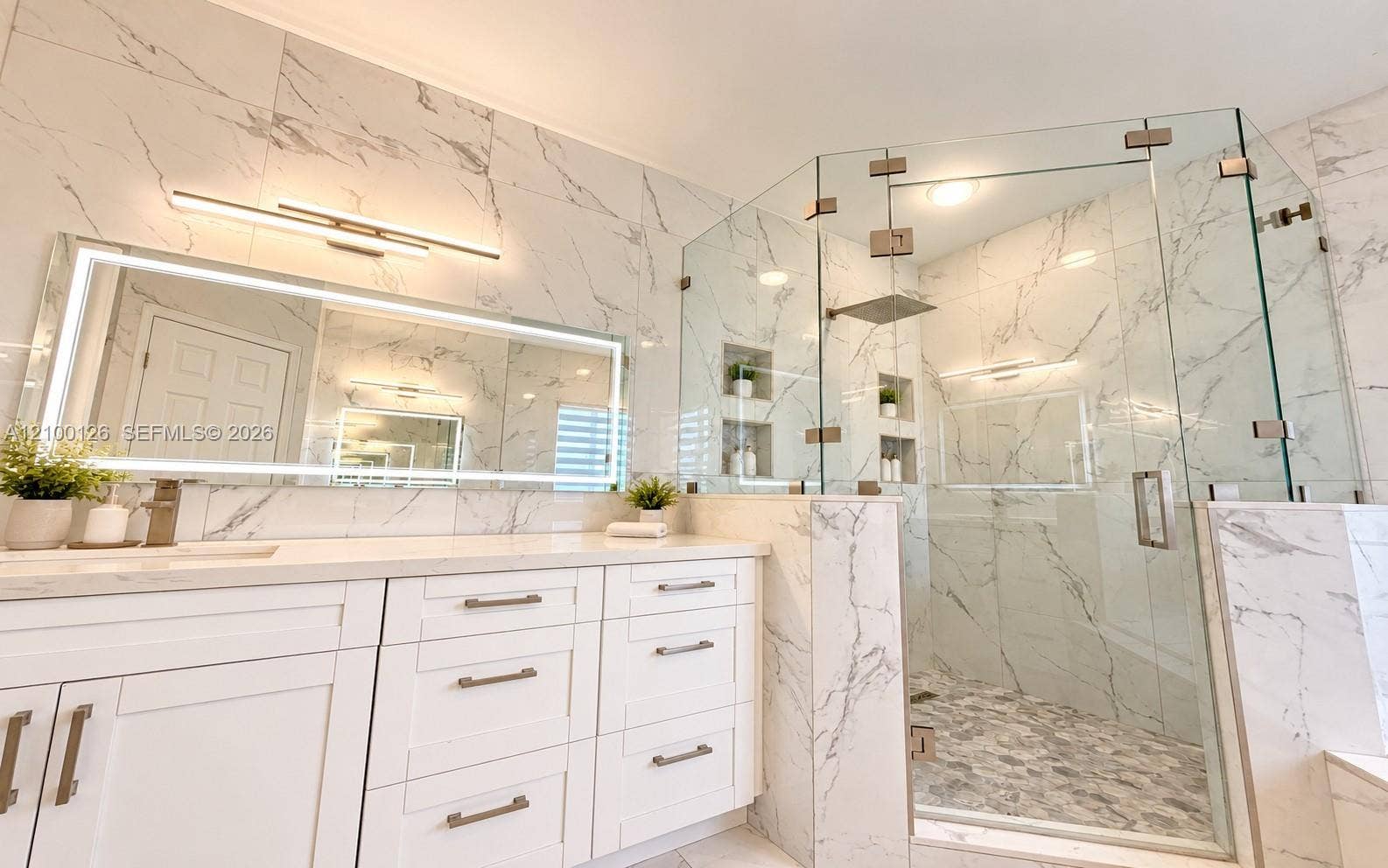 Custom white cabinetry with light-toned countertops, large-format stone-patterned wall tile, frameless glass-enclosed shower with a rainfall showerhead, integrated LED-backlit vanity mirror, and recessed wall shelving