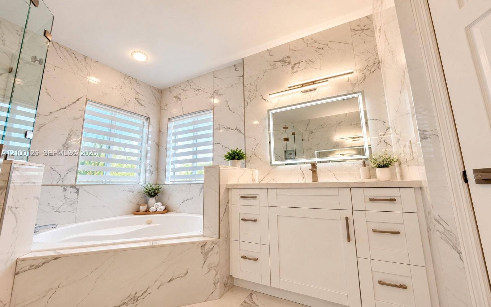 Floor-to-ceiling veined stone tile, white shaker-style vanity with brass-finish hardware, built-in soaking tub, backlit vanity mirror, and glass-enclosed shower