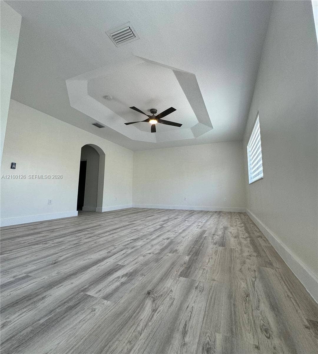 Tray ceiling with a central multi-blade ceiling fan, wood-finish flooring, arched entryway, and dual-layered window shades