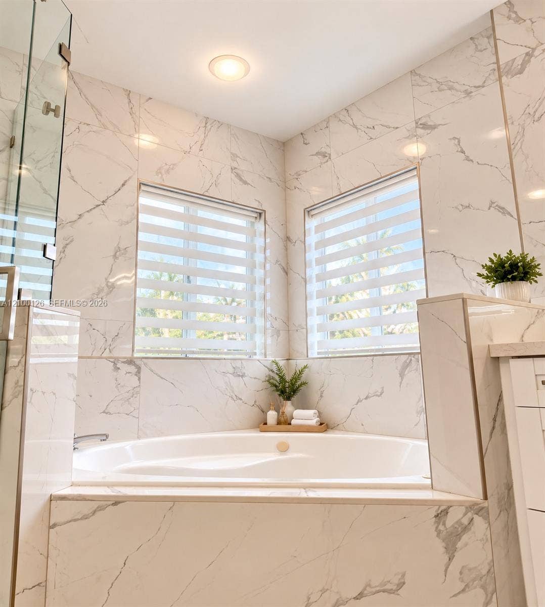 Corner soaking tub featuring full-height large-format tile surround, dual windows, and recessed lighting