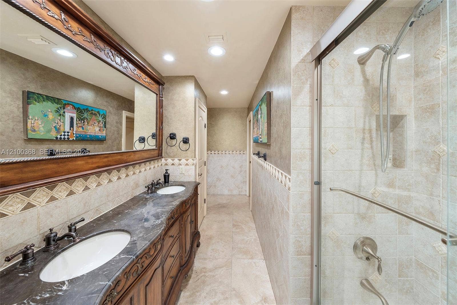 Double vanity with stone countertop, dual undermount sinks, intricate wood-finish cabinetry, a large framed vanity mirror, and a tiled walk-in shower with glass enclosure