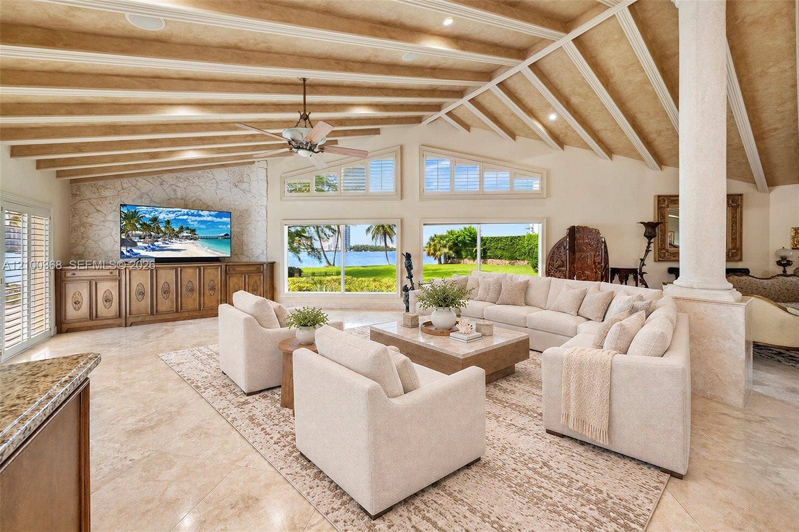 Vaulted beamed ceiling featuring recessed lighting, stone-textured accent wall with integrated cabinetry, polished stone flooring, and expansive windows showcasing a waterfront backdrop
