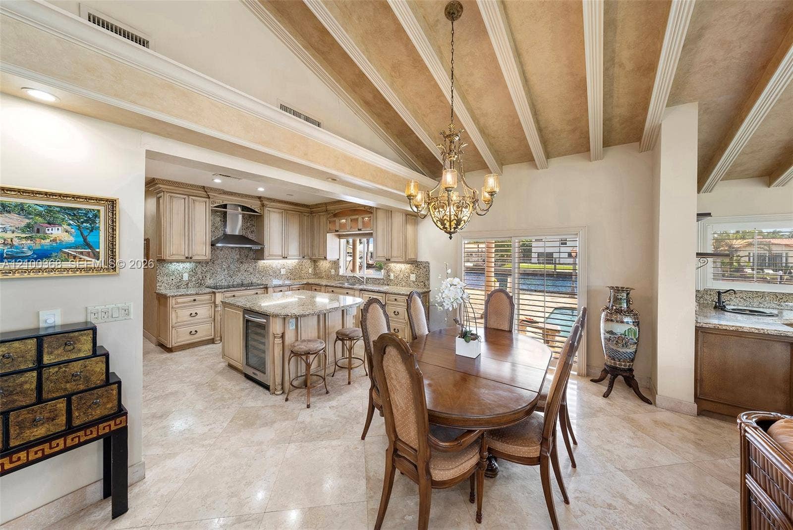 Vaulted ceilings with decorative exposed beams, polished stone flooring, central kitchen island with integrated wine storage, and ornate multi-arm chandelier
